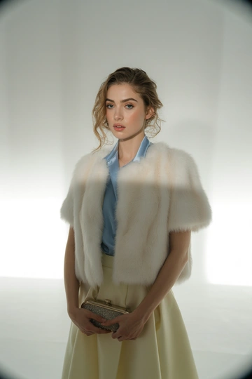 Женщина в студии на белом фоне в элегантной белой шубе с коротким рукавом, под шубой светло-голубая шелковая блуза и узкая кремовая юбка по колено. Стоит в слегка расслабленной позе, смотрит прямо в камеру, гламурный макияж, волосы распущены легкими волнами, в руках миниатюрный клатч.