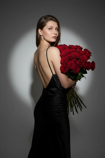  Three-quarter back portrait of a woman holding a large bouquet of deep red roses, head turned three-quarters to the left looking directly at the camera with a neutral expression, eye-level angle, dynamic off-center composition with the bouquet positioned to the right, wearing a black satin slip dress with thin straps and a deep open back exposing the shoulder blades, against a seamless medium gray background in a professional photo studio, dramatic spotlight from the front-top left at a 45-degree angle creating a circular glow in the background and strong halo lighting along the hair and edges of the dress, hard shadows falling to the lower right corner shaping the silhouette, intense and cinematic atmosphere, shot on an 85mm portrait lens with a shallow depth of field, photorealistic high fashion editorial style with visible satin sheen and individual rose petal veins, highly detailed folds fabrics and strands of hair, a limited color palette dominated by deep blacks, rich reds and neutral grays, 8K resolution.