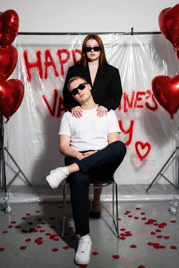 Фотореалистичная редакционная фотосессия пары ко Дню святого Валентина в студии. Фотозона как на референсе: белая студия с прозрачной плёнкой, натянутой на стойках. На плёнке — красные граффити-надписи "Happy Valentine's Day" и сердца в стиле спрей-арта, создающие дерзкий и контрастный фон. По бокам — студийные стойки и красные воздушные шары в форме сердца, на полу разбросаны красные лепестки роз. Атмосфера — современная, fashion, с лёгким арт-хаосом.

Поза и композиция: Крупный план до бедер, мужчина сидит на стуле по центру кадра в позе нога на ногу, уверенная расслабленная поза, его голова немного наклонена в сторону. Девушка стоит за ним, близко, её руки лежат на его плечах или слегка касаются шеи. Между парой ощущение близости и доминирующей, уверенной энергии девушки.
Одежда:
— Мужчина: белая футболка без принтов, тёмные брюки, белые кроссовки. Тёмные очки как fashion-акцент.
— Девушка: чёрный пиджак свободной посадки. Тёмные очки как fashion-акцент.
Выражения лиц: мужчина смотрит в камеру уверенно и спокойно. Девушка смотрит в камеру или слегка сверху вниз, с холодной, контролируемой эмоцией.
Свет: жёсткий студийный свет с акцентом на лица и текстуры, чёткие тени, высокий контраст.
Композиция: вертикальный кадр, пара по центру, фон полностью читается.
Стиль: фотореализм, модная редакционная съёмка, высокая детализация, современный fashion-editorial.