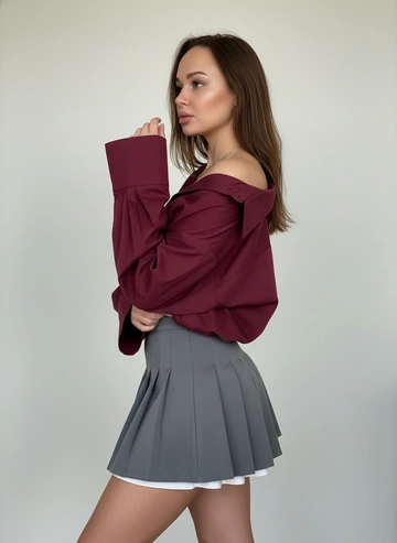 Фото студийное. Фон замыт, на заднем фоне светлая стена. Акцент на девушке.
Не меняя внешности и черты лица.  Девушка снята в профиль по бёдра , поза немного повернута влево: корпус прямой, одна рука согнута у лица, локоть приподнят, плечо обнажено. На ней свободная бордовая (цвет глубокий бордовый) рубашка большого размера с длинными объёмными рукавами и широкими манжетами; рубашка заправлена в пояс юбки так, что одна сторона воротника сдвинута и плечо открытo, создавая небрежно-элегантный эффект. Юбка короткая, плиссированная, трапециевидного кроя серого оттенка; по низу видна белая подкладка, который немного выглядывает. Кожа  загорелого тона; силуэт подчёркнут талией и расклешённой юбкой.
Волосы распущены у лица,видна золотистая  шейная  цепочка.