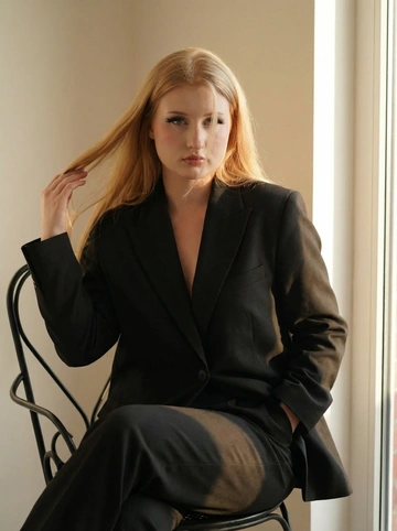   Девушка в студии сидит на чёрном стуле железном, она в чёрном костюме классическом оверсайз, ткань плотная 
Фото по пояс 
Одна рука в кармане 
Другой она поправляет волосы перекидывая их с одной стороны. Пару прядок волос падают на лицо. Сбоку светит солнечный свет частично падает на девушку 
Ресницы пышные 
Кожа идеальная