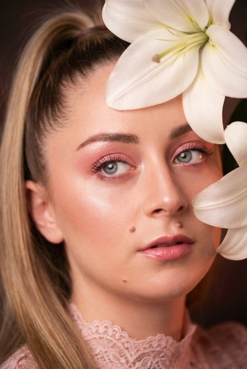   
Крупный план лица женщины, выполненный в студийной обстановке с мягким рассеянным светом. Стиль фотографии - гламурная бьюти-съемка. Цветовая гамма теплая, с преобладанием розовых и персиковых оттенков. Фон размытый, темный, акцент сделан на лице.

Женщина смотрит вверх и немного вбок. Ее лицо обрамлено крупными лепестками белых лилий цветка, частично закрывающими лоб и щеку. Одежда не видна полностью, но заметен фрагмент кружевного розового воротника. На лице присутствует макияж в розовых тонах, с акцентом на глаза и губы. Кожа имеет сияющий эффект.

