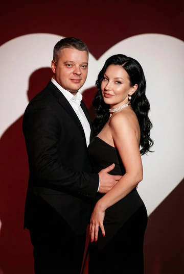 Фотореалистичный редакционный крупный портрет по пояс для пары по референсным фото. Мужчина и женщина позируют вместе, стоят близко. Мужчина одной рукой приобнимает женщину за талию. Фон: тёмно-красный студийный задник, большое белое сердце, проецируемое на стену. Сохранить внешность обоих: реальные черты лица, естественная текстура кожи, реальный цвет и длина волос, без ретуши и изменений. Женщина: облегающее чёрное вечернее платье без бретелей с небольшим разрезом, жемчужный чокер, жемчужные серьги, красный маникюр. Поза в три четверти, спина слегка выгнута, одна рука на поясе или касается руки мужчины. Лёгкая улыбка, взгляд в камеру. Мужчина: чёрный классический костюм, белая рубашка. Прямая осанка, уверенная поза. Взгляд в камеру, легкая улыбка. Свет: драматическое боковое студийное освещение, высокий контраст, чёткие тени на фоне. Композиция: вертикальный кадр по пояс, пара в центре, негативное пространство вокруг сердца. Стиль: фотореализм, модная редакционная съёмка, кинематографическая цветокоррекция (тёплые красные, глубокие чёрные), высокая детализация. Голливудская волна, пробор лёгкий боковой. Верхняя зона чуть приподнята у корней, концы — оформленные волной. 