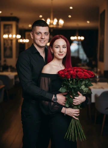 Сохраняя полностью черты лица. Мужчина и женщина в ресторане на фоне столиков.Будто их кто-то фотографирует.Оба смотрят в камеру Мужчина стоит сзади и обнимает женщину за талию.На нем черная классическая рубашка и черные брюки.На руке часы.Женщина стоит в черном вечернем платье с открытыми плечами и длинными рукавами.На ней украшения.В руках будет из 21 красных роз.Волосы распущены по плечам.Оба улыбаются