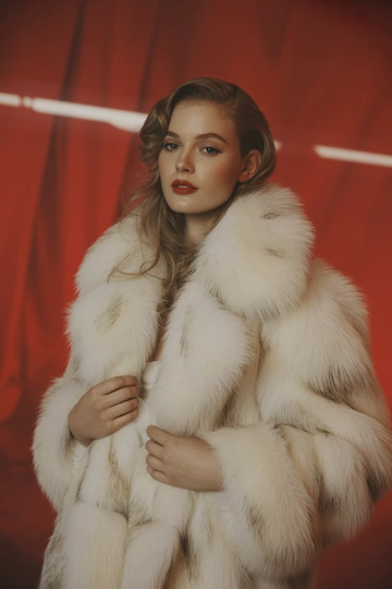 Женщина в студии на красном фоне в роскошной белой шубе с крупным воротником из длинного мягкого меха, средний план, стоит лицом к камере и уверенно смотрит прямо, распущенные волосы, гламурный макияж, атмосфера съемки для винтажного модного ретро-журнала.
