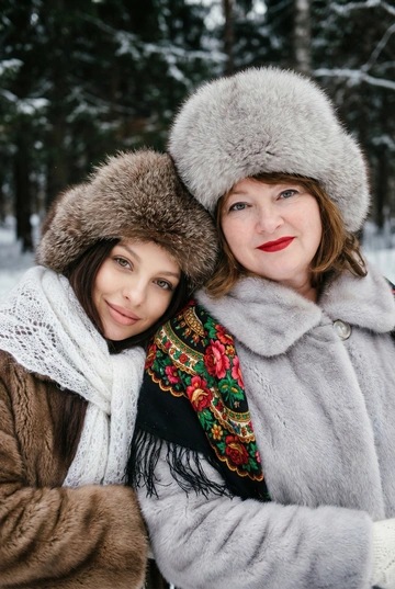 Создай кадр, который представляет собой теплый, уютный, слегка ностальгический зимний портрет в фэшн/фолк-стиле. Атмосфера спокойная, элегантная, дружеская близость двух моделей; сочетание высокой моды (мех, профессиональная ретушь) и фольклорной эстетики (вышитый шарф). Композиция и позирование: Форма кадра: портретный, вертикальный (соотношение примерно 3:4). Расположение: две женщины в центре кадра, тела и головы видны на груди; правая модель немного ближе к камере. Позы: девушки стоят близко друг к другу, левая модель слегка опирается на правую; плечи слегка повернуты к камере. Взаимодействие: мягкая улыбка, закрытое выражение лица, взгляд в камеру; дружелюбный, уверенный тон. Лица и макияж: Кожа: гладкая, естественная текстура, легкий эффект сияния. Глаза: аккуратные блики – оживляют взгляд; нейтральный/теплый макияж теней. Брови: естественная форма, слегка очерчены. Губы: у правой модели насыщенный матовый красный (центральный акцент), у левой – нежно-розовый нюдовый. Волосы: свободные пряди вокруг лица, несколько прядей с легкой волной для естественности. Шапки: большие пушистые меховые шапки с очень четко выраженной текстурой меха; справа – светло-серый мех, слева – коричневый мех. Форма: высокие, округлые, плотно сидят на голове, частично закрывают лоб – создают силуэт и рамку для лица. Верхняя одежда и текстуры: меховые пальто: густой натуральный мех у левой модели коричневый, у правой модели – светло-серый тон с видимым направлением волосков; богатая текстура и объем. У девушки справа на правом плече маленький платок с традиционным русским орнаментом – яркие цветы и листья, на фоне глубокого красного цвета с золотисто-терракотовыми, зелеными деталями; Тонкая бахрома ниспадает на ее плечо. У девушки слева на левом плече небольшой вязаный белый шарф с крупным ажурным узором, что добавляет контраста в текстуре и цвете. Окружение и фон: Лес: темные хвойные деревья, вертикальные силуэты стволов; снег на земле/вдали — размытый фон (боке).