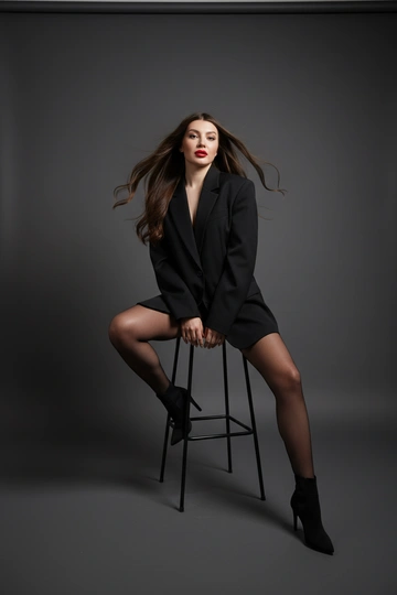 Женщина позирует на высоком стуле в фотостудии. Мягкое профессиональное студийное освещение: верхне-фронтальный ключевой свет, равномерно освещающий лицо и фигуру, без жёстких теней. Свет подчёркивает объём, складки и прозрачность ткани, создаёт чистый, пластичный и дорогой визуальный эффект. Фон тёмно-серый с плавным градиентом, без отвлекающих деталей.. На ней оверсайз пиджак черного цвета, капроновые черные колготки.. У женщины профессиональная укладка - мягкие глянцевые волны.. Волосы слегка развеваются, создавая ощущение движения. Макияж журнальный: ровный сияющий тон кожи, подчёркнутые скулы, деликатный румянец, чётко очерченные губы с красной помадой. Высокая мода, журнальный стиль, минимализм. Женщина сидит на стуле, одна нога согнута и поставлена на нижнюю перекладину стула, вторая опущена на пол. Локоть опирается на поднятое колено, другая рука свободно лежит на бедре. Голова слегка наклонена в камеру.