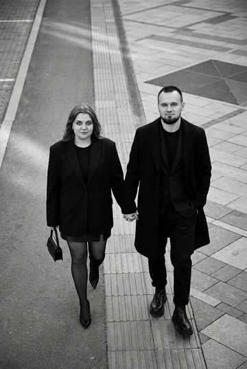 Черно-белая художественная фотография, ультрареализм, 8K, профессиональная съёмка; (оба): в кадре мужчина и женщина; каждый персонаж жёстко привязан к своему загруженному референсу, лица используются напрямую, без генерации, усреднения, подмены или смешения. Вид сверху под углом. Они идут по городской улице, держась за руки; шаг в движении, эффект «пойманного кадра». Смотрим вверх в камеру, выражение спокойное, уверенное, отстранённое.
Полностью чёрные образы modern classic: женщина — оверсайз-пиджак, короткое платье/юбка, плотные колготки, остроносые каблуки, маленькая сумка; мужчина — длинное пальто, свободный костюм, массивные ботинки.
Фон: асфальт, плитка, тактильная дорожка, геометрия.
Контрастный свет, мягкие тени, высокая детализация тканей.
Стиль: black & white, cinematic, high fashion, editorial, minimalism, street photography.
Качество: ultra high resolution, sharp focus, film grain, без ретуши.