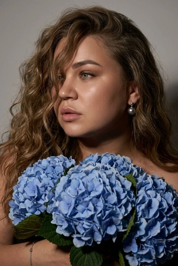 Создайте ультрареалистичный портрет, не меняя черты лица, крупным планом 3/4, с мягкой, освещённой кожей, атласной отделкой и естественным блеском, где женщина изображена в профиле. Спокойное и чувственное выражение лица, губы слегка полуоткрыты. Она носит серебряные серьги-шары и простой серебряный браслет на запястье.
Фон нейтральный, в светло-сером тоне, с тёмным, целенаправленным низким угловым освещением, которое подчёркивает контур лица, текстуру кожи и объём волос. Стиль редактирования — высокая мода, чёткий фокус 8K, реалистичная текстура, изысканная и элегантная атмосфера.


Длинные волнистые волосы покрывают часть лица объёмными локонами, выпадающими на лицо и плечо. 

Цветы: Держит в руках огромный букет голубых гортензий