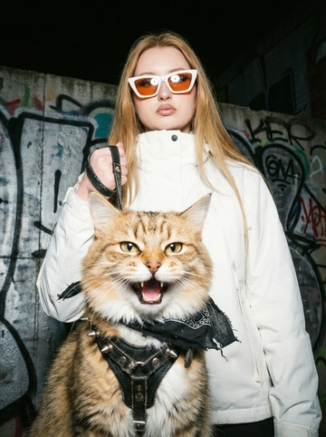         Ночная редакционная уличная портретная съёмка — кадр с крайне низкого угла, с точки зрения «с позиции червя». На переднем плане — крупный план разъярённой кошки. На ней поношенная чёрная кожаная сбруя с тусклыми металлическими шипами и маленькая потрёпанная чёрная бандана, туго завязанная под подбородком. Пасть кота раскрыта в оскале, взгляд резкий и вызывающий.

За ним, занимая большую часть кадра, неподвижно стоит молодая женщина свирепая и стильная. Она небрежно, с показным доминированием держит в одной руке обе сбруи. 

На женщине большие угловатые солнцезащитные белые очки с оранжевыми линзами, в которых отражается резкий прямой свет. На ней яркая белая куртка в стиле gorpcore. Фон — стена с граффити.

Снимок сделан на плёнку Kodak Portra 400 с прямой вспышкой. Изображение выглядит менее привлекательно: высокая контрастность текстур, грубые тусклые полутона, видимая зернистость, лёгкая гало‑эффекта в ярких участках и сильное виньетирование объектива. Формат кадра — 9:16 (вертикальный), что создаёт эффект погружения и передаёт естественную уличную атмосферу.
