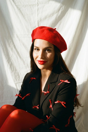 Фотореалистичная редакционная фотосессия женщины в студии. Белый драпированный задник из ткани, напоминающей простыню, с мягкими складками и естественными заломами. Образ: чёрный пиджак свободной посадки с красными декоративными бантиками, красный берет, красные плотные колготки. Макияж во французком стиле с акцентом на красные губы. Сохранить реальную внешность этой женщины, естественную текстуру кожи, без искажений. Стиль съёмки: плёночная фотография, тёплая цветокоррекция, мягкий контраст, лёгкое зерно. Свет естественный, с солнечными бликами и мягкими тенями. Фотореализм, высокая детализация, модная редакционная съёмка. Крупный художественный портрет. Камера близко, лёгкий поворот головы. Одна часть лица в мягкой тени, другая — в солнечном свете. Плёночное зерно, тёплый тон, акцент на глаза и красные губы. Мягкая улыбка.