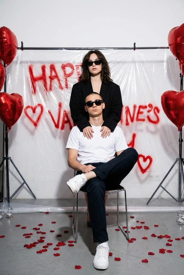 Фотореалистичная редакционная фотосессия пары ко Дню святого Валентина в студии. Фотозона как на референсе: белая студия с прозрачной плёнкой, натянутой на стойках. На плёнке — красные граффити-надписи "Happy Valentine's Day" и сердца в стиле спрей-арта, создающие дерзкий и контрастный фон. По бокам — студийные стойки и красные воздушные шары в форме сердца, на полу разбросаны красные лепестки роз. Атмосфера — современная, fashion, с лёгким арт-хаосом.

Поза и композиция: Крупный план до бедер, мужчина сидит на стуле по центру кадра в позе нога на ногу, уверенная расслабленная поза, его голова немного наклонена в сторону. Девушка стоит за ним, близко, её руки лежат на его плечах или слегка касаются шеи. Между парой ощущение близости и доминирующей, уверенной энергии девушки.
Одежда:
— Мужчина: белая футболка без принтов, тёмные брюки, белые кроссовки. Тёмные очки как fashion-акцент.
— Девушка: чёрный пиджак свободной посадки. Тёмные очки как fashion-акцент.
Выражения лиц: мужчина смотрит в камеру уверенно и спокойно. Девушка смотрит в камеру или слегка сверху вниз, с холодной, контролируемой эмоцией.
Свет: жёсткий студийный свет с акцентом на лица и текстуры, чёткие тени, высокий контраст.
Композиция: вертикальный кадр, пара по центру, фон полностью читается.
Стиль: фотореализм, модная редакционная съёмка, высокая детализация, современный fashion-editorial.