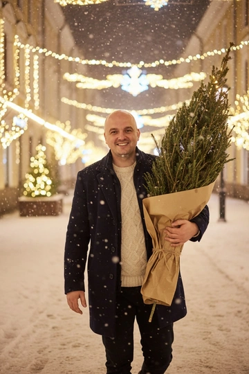 На фоне заснеженного вечернего города, укутанного рождественскими гирляндами и украшениями, падают крупные хлопья снега. Он стоит в темно-синем пальто и светлом уютном свитере. В теплых, мягких тонах, фото передает движение момента, а его искренняя улыбка добавляет ощущения уюта и волшебства зимнего вечера. Идет, неся в руках большую елку, завернутую в коричневую бумагу. Улыбается.