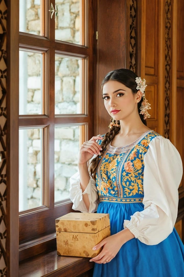 Женщина в длинном синем сарафане с золотой вышивкой и белой рубахой стоит у резного деревянного окна, держит берестяную шкатулку. Она слегка наклоняется вперёд, внимательно и уверенно смотрит прямо в камеру. На заднем плане — резьба, тёплый свет, акцент на её лице и аутентичном костюме.