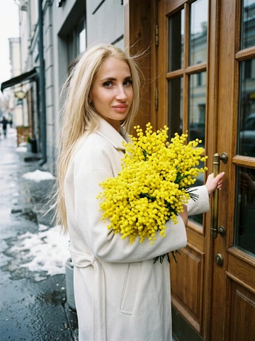 Сохранить черты лица и внешность, возраст, натуральный тон без ретуши. Средне-крупный план (от талии вверх), камера слева от женщины, лицо занимает не менее 35% кадра. Женщина стоит у входа в городское кафе с деревянной дверью и стеклянными вставками, корпус слегка развернут в 3/4 к камере. Лицом к двери, одной рукой держит ручку закрытой двери, другой прижимает к груди огромный пышный букет ярко-жёлтой мимозы. На ней элегантное светлое пальто (молочный оттенок), свободный крой. Волосы как на исходном фото слегка взъерошены ветром Мягкое выражение лица, лёгкая улыбка, взгляд в камеру. Атмосфера начала весны: тающий снег по краям улицы, влажный асфальт, мягкий рассеянный дневной свет, лёгкая прохлада в воздухе. Цветовая палитра нейтральная с ярким акцентом жёлтой мимозы. Фотореализм, натуральная текстура ткани и цветов, без HDR, без пластика, мягкий контраст, 35mm look.