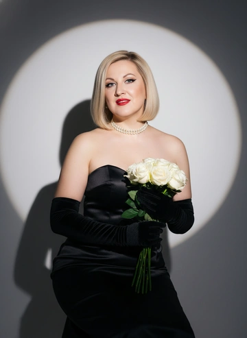 Черты лица строго как на фото. Максимальная схожесть
Женщина позирует в фотостудии. На женщине Черное сатиновое платье без бретелей, подчёркивающее силуэт, жемчужный чокер на шее, черные бархатные перчатки выше локтя. Прическа - удлиненный боб, волосы светлые.  Макияж - четкие черные стрелочки на глазах, губы окрашены в ярко красный цвет. Реалистичная сияюзая текстура кожи, профессиональный макияж. Один свет прожектора, фон - градиент от темно-серого к белому с круглым световым пятном, чёткая тень за спиной. Высокое разрешение, ультрареалистичный, 8K, стиль студийной модной фотографии. Крупный план. Она сидит, держа в руках букет белых роз. На лице спокойная улыбка.Фон нейтральный, свет студийный, мягко выделяющий силуэт.