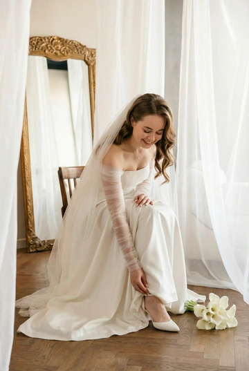 Современная свадебная фотосессия в светлой студии как на референсе: белые занавески, паркет, зеркало в золотой раме, мягкий естественный дневной свет. Эстетика editorial wedding и lifestyle. Не меняй внешность, комплекцию, цвет волос и возраст людей на первых фото. Жених в тёмном классическом костюме с бутоньеркой, темный галстук-бабочка, невеста в белом платье как на референсе, свадебный макияж. У женщины профессиональная укладка - мягкие глянцевые волны.. На голове у женщины лёгкая прозрачная свадебная фата, ниспадающая на плечи и спину. На шее у невесты тонкая цепочка из белого золота и мальенький брллиантовый кулон. Акцент на близость, прикосновения и живые эмоции. Кадр выглядит естественно, без жёсткого позирования. Реалистичное сходство, профессиональная свадебная фотография. Теплая цветовая гамма. В руках у невесты букет из белых калл. Жених такого же роста, как невеста .  Портрет невесты. Невеста сидит и поправляет туфлю или ремешок, платье свободно драпируется. Свет мягкий, подчёркивает форму и ткань.