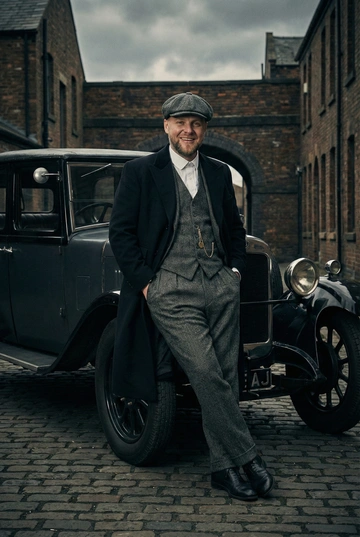 Кинематографичная мужская фотосессия в эстетике британской гангстерской драмы 1920-х годов. Мужчина в классическом костюме-тройке из твида как на референсе: пиджак, жилет, брюки, светлая рубашка, галстук. Если у мужчины на референсе есть волосы, то сделай ему аккуратная классическая стрижка с короткими боками, естественная укладка, цвет волос сохранён. Если у мужчины волос нет, то прическу не трогай. Образ строгий, собранный, харизматичный. Выражение лица сдержанное, уверенное. Свет драматичный, направленный, с глубокими тенями. Цветокоррекция приглушённая, кинематографичная. Фокус на человеке, профессиональное качество. Мужчина стоит, опираясь спиной на винтажный автомобиль 1920-х годов. Он в костюме-тройке и длинном пальто, на голове твидовая кепка. Руки в карманах брюк. Фон — кирпичное здание и арка. Пасмурный дневной свет, кинематографичная улица.