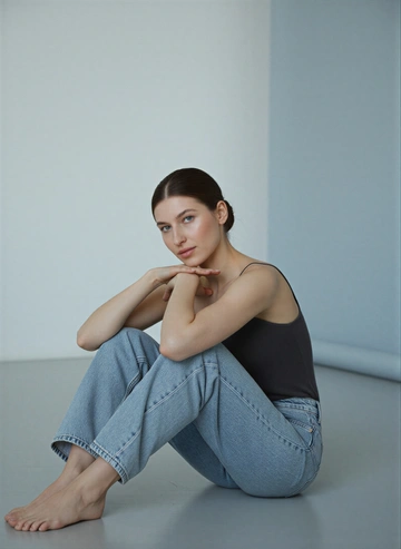 Лицо не меняй, строго как на исходном фото. Женщина сидит на полу в светлой, минималистичной студии. Это кадр из модной фотосессии с акцентом на естественную красоту и спокойную атмосферу. Цветовая гамма нейтральная, с преобладанием светлых оттенков серого и голубого.

На женщине надет темный топ на тонких бретельках и светлые джинсы свободного кроя. Она сидит, подтянув колени к груди, и опирается подбородком на руки, лежащие на коленях. Взгляд направлен прямо в камеру. Поза расслабленная и непринужденная. Волосы собраны в гладкий пучок. Лёгкий макияж с сияющий хайлайтером.
