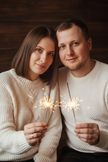 Используй ДВА загруженных фото: мужчина и женщина. Не изменяй лица исходных фото (черты, пропорции, возраст, мимику). Сохраняй естественные телосложения. Очки и аксессуары сохраняй только если есть на исходниках. Делай реалистичную перспективу и тени, избегай «пластиковости». Не меняя внешности людей с фото, запечатлена пара в теплой атмосфере. Женщина слегка наклонила голову к мужчине, лица близко, оба смотрят в объектив с лёгкой улыбкой, сохраняя естественные черты. Женщина в уютном белом свитере с выразительной вязкой, видна текстура ткани при мягком рассеянном свете. Мужчина в белом свитере с мелким фактурным узором, облегающем фигуру. Освещение — мягкий вечерний свет с огненными бликами от бенгальских огней в их руках, свет играет на лицах и одежде, создавая тёплые тени и подсветку. Размытый тёмный фон с деревянной структурой, придающий уют и загадочность. Композиция фокусируется на лицах и бликах огней, создавая праздник и близость. Тёплая золотая палитра, мягкие тени, кинематографичное, фотореалистичное, высокая детализация, 4K.