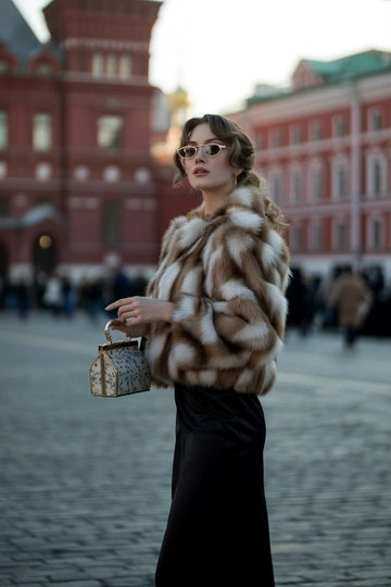 Женщина на фоне Красной площади в роскошной соболиной шубе, под шубой элегантное черное платье миди. Стоит в пол-оборота, смотрит прямо в камеру. На лице – гламурный макияж, волосы распущены, в руках маленькая лакированная сумочка, образ дополняют ретро солнечные очки.