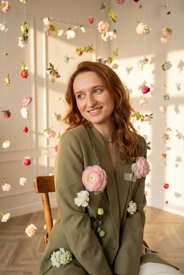 Студийная фотосессия этой женщины в интерьерной студии с подвешенными цветами. Используй приложенное изображение студии как точный визуальный референс локации: светлые стены, мягкий дневной свет, множество живых цветов и бутонов, подвешенных на тонких нитях, создающих ощущение парящего цветочного поля. Цветы расположены как на фоне, так и в переднем плане. Образ: на женщине один зеленый пиджак с цветами как на референсе. Волосы распущены, уложены в легкие волны.. Макияж натуральный, свежий, с акцентом на кожу и мягкие черты лица. Цветокоррекция теплая, пастельная, golden hour. Настроение фотосессии — естественность, нежность, интимность, ощущение тишины и спокойного момента. Профессиональная съёмка, реалистичное качество, без искажений лица и пропорций. Цвета пастельные, с тёплым солнечным подтоном. Лёгкий эффект плёночности, без потери детализации лица. Крупный план. Женщина сидит на деревянном стуле нога на ногу и слегка улыбается, взгляд направлен в сторону, как будто она реагирует на что-то вне кадра. Голова немного наклонена. Настроение живое и естественное.  Пиджак спущен с одного плеча, обнажая его.