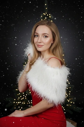 Праздничная студийная фотосессия. Женщина одета в красное вечернее платье на тонких бретелях, с белой пушистой накидкой из перьев. Фон — градиент от черного к тёмно-серому, мягкий тёплый направленный свет сбоку, лёгкие светлые частицы в воздухе создают эффект падающего снега. Позади стоит минималистичная рождественская ель, украшенной только гирляндой тёплого света. Атмосфера — элегантная, глянцевая, как для зимней рекламной кампании или обложки журнала. Реалистичный стиль, чистая кожа, аккуратный макияж, акцент на блеске украшений и фактуре перьев. Волосы длинные, блестящие и густые. Крупный план лица. Женщина смотрит прямо в камеру, лёгкая улыбка, глаза блестят. Накидка мягко касаются щеки, создавая интимное и нежное настроение. Свет мягкий, акцент на лице и блеске украшений.