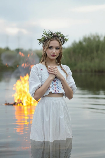 Молодая женщина в белой хлопковой юбке и чуть укороченной рубахе с вышитыми красными и синими узорами, стоит по пояс в воде. Венок из свежих луговых трав украшает её волосы. Она смотрит прямо в камеру, обе ладони прижаты к груди, за спиной мерцает костёр и отражается в воде.