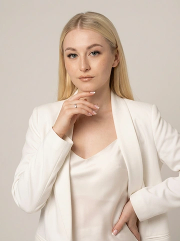   Не менять черты лица. Студийный портрет с мягким освещением. Девушка с нежными чертами лица, цвет волос блонд, длинные прямые блестящие волосы с прямым пробором по середине, волосы собраны за уши. Белоснежная чистая кожа с небольшими веснушками на лице. Белый строгий пиджак кимоно. Под пиджаком надето шелковое платье на брительках молочного оттенка. Лицо в три четверти, прямая осанка, правая рука мягко лежит на бедре, левая рука мягко касается подбородка указательным пальцем, на безымянном пальце серебряное кольцо в минималистичном стиле. Маникюр средней длины, белый французский маникюр. 
Сделать акцент на длинных натуральных ресницах. Макияж нежный нюдовый. Придать небольшой блеск на губах плампером , цвет губ легкий бежевый, ближе к естественному оттенку губ