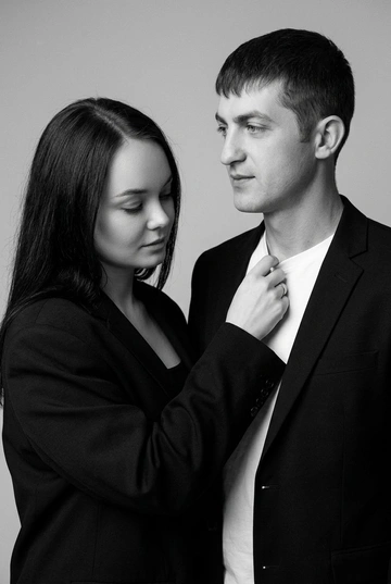 Черно-белый портрет мужчины и женщины в стиле фэшн фотосессии в фотостудии. Не изменяйте лица. Фон нейтральный, однотонный, светло-серый. Атмосфера интимная и спокойная. На переднем плане женщина. Она одета в черный оверсайз пиджак и черный топ. Мужчина одет в в классическом черном пиджаке и белой футболке. Вся композиция выполнена в приглушенной монохромной гамме со студийным освещением, которое подчеркивает текстуру кожи и одежды.  Женщина поправляет рукой его футболку, при этом оба лица хорошо освещены и видны.