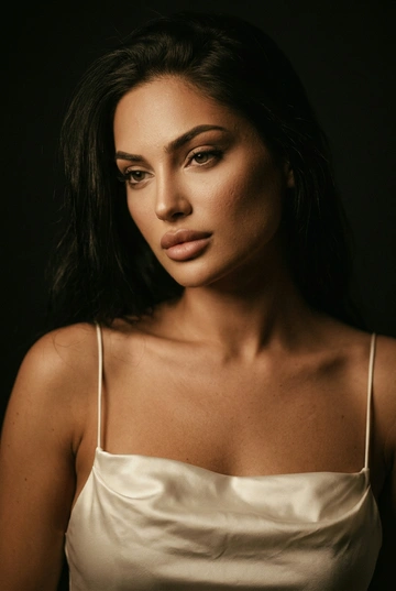 Художественный портрет женщины в полумраке. Камерная атмосфера, тёмный фон без деталей. На ней атласное платье-компбинация нежно-молочного оттенка с тонкими бретелями. Ткань ловит свет, создавая мягкие блики и шелковистую текстуру. Освещение мягкое, направленное, с эффектом старого студийного света, плавные тени, тёплая цветокоррекция, лёгкий винтажный эффект, кинематографичная плёночная зернистость. Реалистичное лицо, естественные пропорции, высокая детализация кожи, профессиональное качество, fine art fashion портрет. Крупный портрет. Голова слегка наклонена, выражение спокойное. Тёплый свет подчёркивает глаза и скулы, винтажная цветовая гамма, эффект плёночной съёмки.