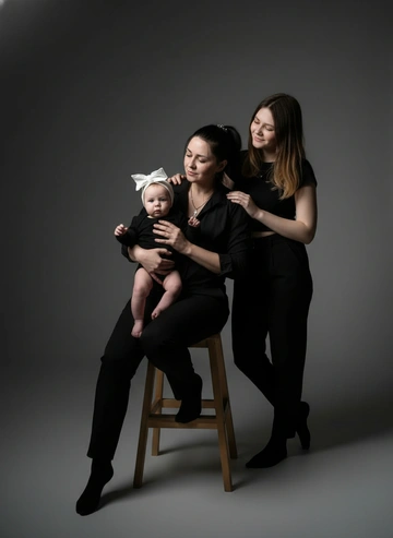 Максимально сохранить черты лиц. Студийная съёмка. В лучах прожектора. Мама сидит на стуле, держит ребёнка на коленях. Девушка стоит сзади, положив руку на плечо маме. Одеты в чёрное
