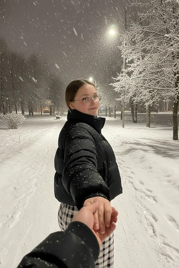 Создай на этом фото романтический и слегка меланхоличный момент, снятый от первого лица (POV) ночью, на фоне зимнего городского парка.
В центре кадра — девушка. Её лицо полностью открыто и хорошо видно. Девушка держит парня за руку и резко оборачивается через плечо, мило улыбается Взгляд направлен прямо в камеру
Одежда и детали:
На девушке объёмная укороченная чёрная куртка, выглядящая тёплой, уютной и современной, идеально подходящей для зимней ночной прогулки.
Взаимодействие:
На переднем плане видна рука парня — это рука человека, который делает снимок. Их пальцы переплетены, что создаёт классический сюжет «follow me» (следуй за мной) и подчёркивает близость пары. Рука парня одета в чёрную куртку с ребристыми манжетами.
Окружающая обстановка:
• Место: ночной заснеженный городской парк
• Пейзаж: справа — деревья, густо покрытые снегом, и уличные фонари с мягким тёплым светом
• Атмосфера: зимняя эстетика — прохладное, немного грустное, но очень уютное настроение. В воздухе видны крупные хлопья падающего снега, создающие ощущение движения и глубины кадра.
Общее впечатление:
Фотография передаёт момент прогулки, наполненной движением, ветром и эмоциональной близостью. Кадр выглядит как стоп-кадр из лайфстайл-блога или короткого видео для Reels/TikTok, где романтичное начало создаёт интригу и ощущение неожиданного поворота.