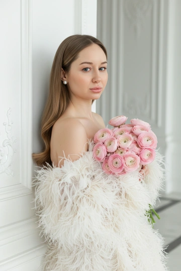 Создай крупный плечевой портрет, не меняя черты лица.Детальный крупный портрет элегантной женщины в эффектном, роскошном, модном платье. Женщина с волнистыми волосами. Одежда представляет собой объемное творение кутюр, полностью сделанное из больших, текстурированных, кремово-белых перьев, уложенного с открытыми плечами, чтобы обнажить декольте  Женщина стоит в динамичной, чувственной позе, прислонившись к белой классическому архитектурной стене. На ней серьги-гвоздики жемчужные . На заднем плане представлен грандиозный, классический интерьер/экстерьер - ярко-белая мраморная или лепная стена. Редакционное освещение, мягкое и рассеянное, подчеркивающее текстуру перьев и кожи.в руках букет. Высокое разрешение, сверхдетализация, модная фотография Букет из нежно-розовых ранункулюсов 