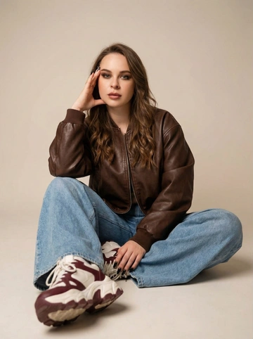   Фото 3:4. Студийный портрет в полный рост молодой женщины в стиле «streetwear chic» и «oversize fashion». Она сидит на полу в расслабленной, но уверенной позе. Фотография выполнена в мягких, теплых тонах с акцентом на текстуры кожи и денима.
Персонаж и внешность:
• Внешность: Девушка с длинными  волосами, уложенными мягкими небрежными волнами. У нее выразительный взгляд, подчеркнутый макияжем в стиле «smoky eyes» в коричневых тонах, и матовые губы.
• Маникюр: Длинные ногти, покрытые черным лаком.
• Одежда: * Объемная кожаная куртка (бомбер) шоколадного или темно-коричневого цвета с матовым блеском.
• Очень широкие светло-голубые джинсы (wide-leg/baggy jeans) из плотного денима, которые создают объемные складки.
• Массивные кроссовки (sneakers) в бело-бордовой цветовой гамме.
• Поза: Девушка сидит, широко расставив ноги, одна рука поднесена к лицу (пальцы касаются виска), локоть опирается на колено.
Окружение и свет:
• Фон: Минималистичная профессиональная студия.Однотонный светло-бежевый или молочный фон без лишних деталей, создающий мягкий градиент.
• Освещение: Мягкий рассеянный свет (softbox), который плавно подчеркивает контуры лица и объем одежды, не создавая жестких теней. Эффект студийной глубины резкости (bokeh) — кроссовки на переднем плане слегка размыты, фокус сосредоточен на лице.
Технические параметры стиля:
• Стиль: Lookbook современной моды, эстетика Pinterest, журнальная съемка.
• Цветовая палитра: Теплая, землистая гамма — шоколадный, бежевый, приглушенный голубой и глубокий бордовый.
• Качество: Сверхвысокая детализация, реалистичная текстура кожи и кожи куртки, 8k, профессиональная ретушь.