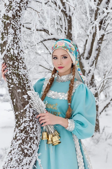 Девушка в бирюзовом сарафане с серебряной отделкой и расписной платочной повязке стоит у изогнутого дерева, покрытого инеем, держит в руках связку колокольчиков, смотрит прямо в камеру, вокруг воздушные снежинки и морозная дымка.