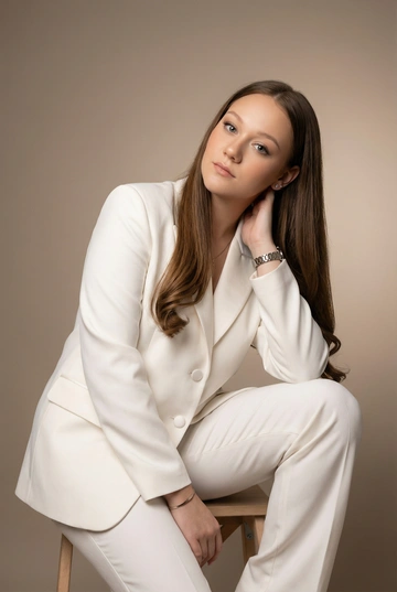 Профессиональная портретная фотосессия для деловой женщины. Женщина одета в белый классический пиджак как на референсе, и белые брюки. Образ элегантный, современный, статусный. Волосы ухоженные, прямые, подкрученные концы, цвет и длина волос сохранёны.. Макияж профессиональный, деловой, с акцентом на свежий тон кожи и аккуратно оформленные глаза. Естественная текстура кожи, губы накрашены помадой естественного цвета. Украшения минималистичные: серьги, часы и тонкий браслет. Фон нейтральный студийный мягкий тёплый градиент, без лишних деталей.. Мягкий рассеянный студийный свет, аккуратные тени, ровная экспозиция. Фокус на лице, четкие черты, профессиональное качество съемки. Настроение — уверенность, спокойствие, экспертность. Кадрирование портретное (по грудь или по пояс), подходит для делового профиля, сайта и LinkedIn. Женщина сидит на стуле, развернутая боком к камере. Корпус слегка наклонен вперед. Правая нога согнута и стоит на полу, левая нога опущена и расслаблена. Левая рука лежит на колене. Правая рука поднята и касается шеи под ухом. Голова наклонена в сторону поднятой руки. Взгляд направлен прямо в камеру. Поза спокойная, элегантная, уверенная.