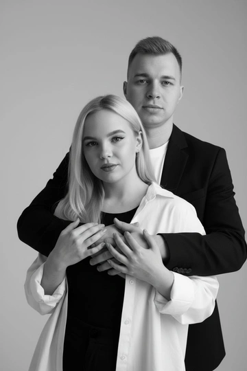 Черно-белый портрет мужчины и женщины в стиле фэшн фотосессии в фотостудии. Не изменяйте лица. Фон нейтральный, однотонный, светло-серый. Атмосфера интимная и спокойная. На переднем плане женщина. Вся композиция выполнена в приглушенной монохромной гамме со студийным освещением, которое подчеркивает текстуру кожи и одежды. Её руки лежат на груди мужчины. Мужчина обнимает женщину сзади, положив свою руку поверх её руки. Она одета в черный топ, белую оверсайз рубашку. Мужчина одет в  классическом черном пиджаке и белой футболке.