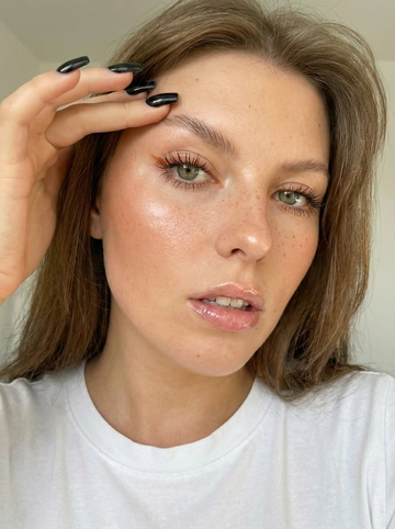     На этой фотографии представлено эстетичное бьюти-селфи молодой женщины. Снимок выполнен в высоком качестве с акцентом на детали макияжа и внешности.
Лицо и макияж:
*   Губы: блеск на губах , Губы слегка приоткрыты, слегка обнажая зубы.
*   Глаза: У девушки яркие глаза. Нанесен макияж с эффектом «лисьего взгляда»: тонкие растушеванные стрелки и длинные, густые, разделенные ресницы, которые удлиняются к внешним уголкам глаз.
Рукой она подтягивает бровь 
Ногти длинные черные  квадратные 
*   Кожа: Выглядит слегка загорелой и сияющей (эффект «dewy skin»). На переносице заметны очаровательные веснушки. На скулах и кончике носа нанесен хайлайтер, создающий мягкие блики.
*   Брови: Натурального оттенка, уложены волосок к волоску, имеют мягкий изгиб.


Одежда и фон:
*   Одежда: Девушка одета в простую базовую белую футболку, что не отвлекает внимания от её лица.
*   Фон: Минималистичный, светлый (почти белый), что создает чистый и современный стиль фотографии.

Ракурс и освещение:
*   Ракурс: Крупный план (портрет), голова полубоком, взгляд направлен прямо в объектив, что создает доверительный и притягательный образ.
*   Освещение: Мягкое, рассеянное дневное освещение. Оно равномерно ложится на лицо, подчеркивая текстуру кожи и блеск волос.

Общее впечатление от фотографии — современный, ухоженный и стильный бьюти-образ, сочетающий в себе естественность (веснушки, мягкие локоны) и драматичный акцент (темные губы).
Черты лица-полностью сохранены как на исходном фото. Фото 3:4