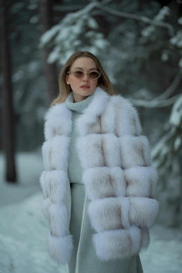 Женщина в заснеженном лесу в роскошной белой шубе средней длины, под ней светло-серое трикотажное платье с высокой горловиной. Стоит в среднем плане, смотрит прямо в камеру, на лице естественный макияж, волосы распущены, атмосферу дополняют ретро солнечные очки и теплые сапоги.