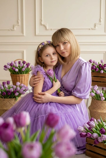 Студийная весенняя фотосессия мамы и дочки в нежной фиолетовой цветовой гамме. Вокруг расставлены корзины и ящики с большим количеством фиолетовых тюльпанов. Мама в нежно-фиолетовом атласном платье с V-вырезом и объемными рукавами, естественный сияющий макияж. Дочка в пышном платье из фиолетового фатина с маленьким цветочным ободком.. Дочери 10 лет. Не меняй прическу матери, она должна быть такой же, как на предоставленном фото.. Светлый минималистичный фон c лепниной. Мягкий рассеянный студийный свет, чистая профессиональная цветокоррекция, высокая детализация, естественная поза. Не менять черты лица, возраст дочери и матери, цвет и длину волос. Атмосфера кадра — нежная, теплая, с акцентом на эмоциях и тактильности. Реалистичное качество, без искажений лица и пропорций. Крупный портретный кадр. Дочка на руках у мамы, держит один тюльпан у груди. Обе смотрят прямо в камеру, спокойное нежное выражение лица. Цветы на переднем плане создают лёгкую глубину.