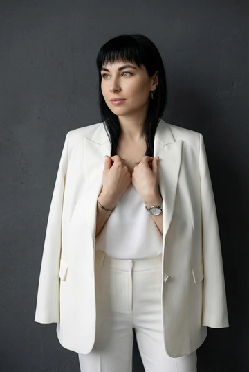 Профессиональная портретная фотосессия для деловой женщины. Женщина одета в белый классический пиджак как на референсе, и белые брюки. Образ элегантный, современный, статусный. Не меняй прическу, она должна быть такой же, как на предоставленном фото.. Макияж профессиональный, деловой, с акцентом на свежий тон кожи и аккуратно оформленные глаза. Естественная текстура кожи, губы накрашены помадой естественного цвета. Украшения минималистичные: серьги, часы и тонкий браслет. Фон – темно-серая фактурная стена.. Мягкий рассеянный студийный свет, аккуратные тени, ровная экспозиция. Фокус на лице, четкие черты, профессиональное качество съемки. Настроение — уверенность, спокойствие, экспертность. Кадрирование портретное (по грудь или по пояс), подходит для делового профиля, сайта и LinkedIn. Портрет по пояс стоя. Женщина стоит прямо, корпус слегка развернут в сторону. Женщина одета в белый топ, пиджак накинут на плечи. Ладони сложены вместе на уровне груди, пальцы расслаблены. Голова повернута в сторону, взгляд направлен вдаль. Поза спокойная, уверенная, с акцентом на осанку.