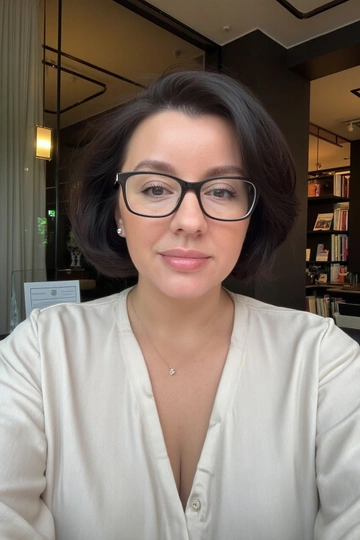 Женщина в светлой шелковой блузке, с объемной аккуратно уложенной прической и легким макияжем, сидит в офисном интерьере с современным дизайном, на фоне книжных полок или рабочего стола с ноутбуком и бумагами. Освещение мягкое и равномерное, подчеркивающее черты лица и текстуру блузки. Поза уверенная, смотрит прямо в камеру, выражение лица деловое и интеллектуальное, демонстрирующее профессионализм и утонченность. Кадр крупный или средний, акцент на лице и верхней части тела.