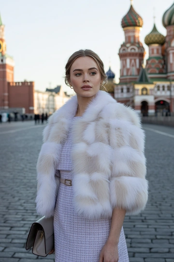 Женщина на фоне Красной площади в короткой белой шубе с пушистым воротником, под шубой светло-лавандовое платье из твида с тонким поясом. Стоит в среднем плане, смотрит прямо в камеру. На лице гламурный макияж, волосы распущены, в руках аккуратная кожаная сумочка.
