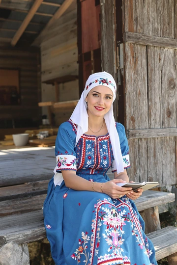 Женщина в голубом сарафане с вышивкой и белой косынке сидит на ступеньках деревянной избы, в руках держит букварь. Она внимательно и доброжелательно смотрит прямо в камеру, на лице мягкая улыбка. Средний план, солнечный день, акцент на традиционном наряде.