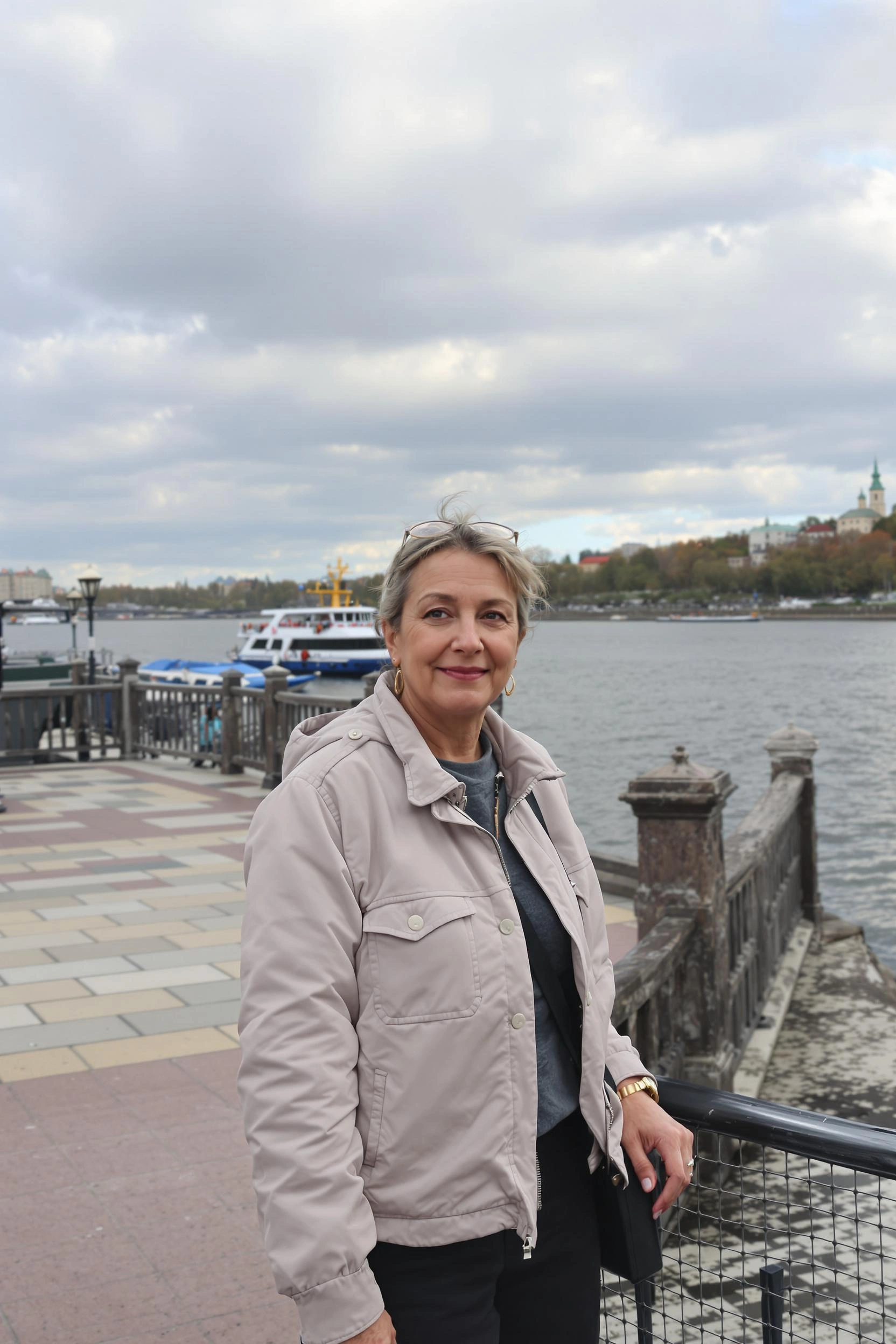 Женщина стоит у деревянных перил набережной, устремив спокойный взгляд в камеру, легкая полуулыбка. За спиной — излучина широкой реки, катер на воде, тротуар из плитки, живописное облачное небо, средний план.