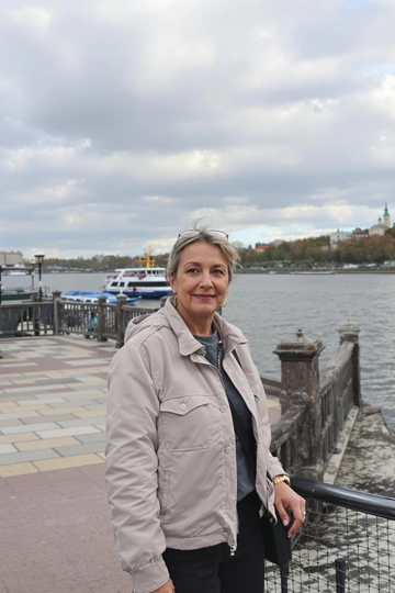 Женщина стоит у деревянных перил набережной, устремив спокойный взгляд в камеру, легкая полуулыбка. За спиной — излучина широкой реки, катер на воде, тротуар из плитки, живописное облачное небо, средний план.
