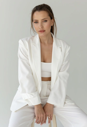Крупный план: женщина позирует на высоком стуле с тонкими ножками в фотостудии цвета белый. На ней костюмный пиджак с широкими плечами белый цвета, застёгнутый на одну пуговицу, и широкие брюки-палаццо белый цвета и кроп-топ в тон. Одежда выполнена из плотной костюмной ткани, держащей форму. Волосы слегка развеваются. Макияж: сияющие скулы, легкие румяна, ровный тон кожи, губы накрашены красной помадой. Высокая мода, журнальный стиль, минимализм. Женщина слегка наклоняется вперёд, локти опираются на колени, кисти рук соединены или свободно лежат. Спина прямая, подбородок немного опущен, взгляд направлен в камеру или в камеру.