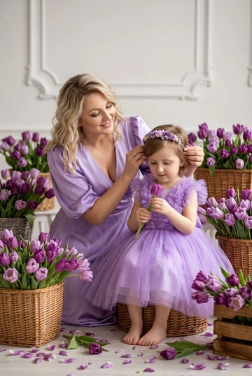 Студийная весенняя фотосессия мамы и дочки в нежной фиолетовой цветовой гамме. Вокруг расставлены корзины и ящики с большим количеством фиолетовых тюльпанов. Мама в нежно-фиолетовом атласном платье с V-вырезом и объемными рукавами, естественный сияющий макияж. Дочка в пышном платье из фиолетового фатина с маленьким цветочным ободком.. Дочери 5 лет. У мамы волосы распущены, уложены в легкие волны.. Светлый минималистичный фон c лепниной. Мягкий рассеянный студийный свет, чистая профессиональная цветокоррекция, высокая детализация, естественная поза. Не менять черты лица, возраст дочери и матери, цвет и длину волос. Атмосфера кадра — нежная, теплая, с акцентом на эмоциях и тактильности. Реалистичное качество, без искажений лица и пропорций. Мама слегка наклоняется к дочке, поправляя ей ободок. Дочка держит один тюльпан и рассматривает его. Камера чуть ниже уровня глаз ребенка, теплый живой момент.