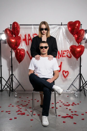 Фотореалистичная редакционная фотосессия пары ко Дню святого Валентина в студии. Фотозона как на референсе: белая студия с прозрачной плёнкой, натянутой на стойках. На плёнке — красные граффити-надписи "Happy Valentine's Day" и сердца в стиле спрей-арта, создающие дерзкий и контрастный фон. По бокам — студийные стойки и красные воздушные шары в форме сердца, на полу разбросаны красные лепестки роз. Атмосфера — современная, fashion, с лёгким арт-хаосом.

Поза и композиция: Крупный план до бедер, мужчина сидит на стуле по центру кадра в позе нога на ногу, уверенная расслабленная поза, его голова немного наклонена в сторону. Девушка стоит за ним, близко, её руки лежат на его плечах или слегка касаются шеи. Между парой ощущение близости и доминирующей, уверенной энергии девушки.
Одежда:
— Мужчина: белая футболка без принтов, тёмные брюки, белые кроссовки. Тёмные очки как fashion-акцент.
— Девушка: чёрный пиджак свободной посадки. Тёмные очки как fashion-акцент.
Выражения лиц: мужчина смотрит в камеру уверенно и спокойно. Девушка смотрит в камеру или слегка сверху вниз, с холодной, контролируемой эмоцией.
Свет: жёсткий студийный свет с акцентом на лица и текстуры, чёткие тени, высокий контраст.
Композиция: вертикальный кадр, пара по центру, фон полностью читается.
Стиль: фотореализм, модная редакционная съёмка, высокая детализация, современный fashion-editorial.